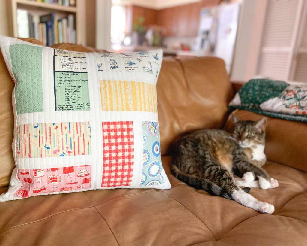 Finished Piece of Cake Pillow made with bright geometric fabric rectangles and white borders, styled on a modern sofa with a lazy cat.